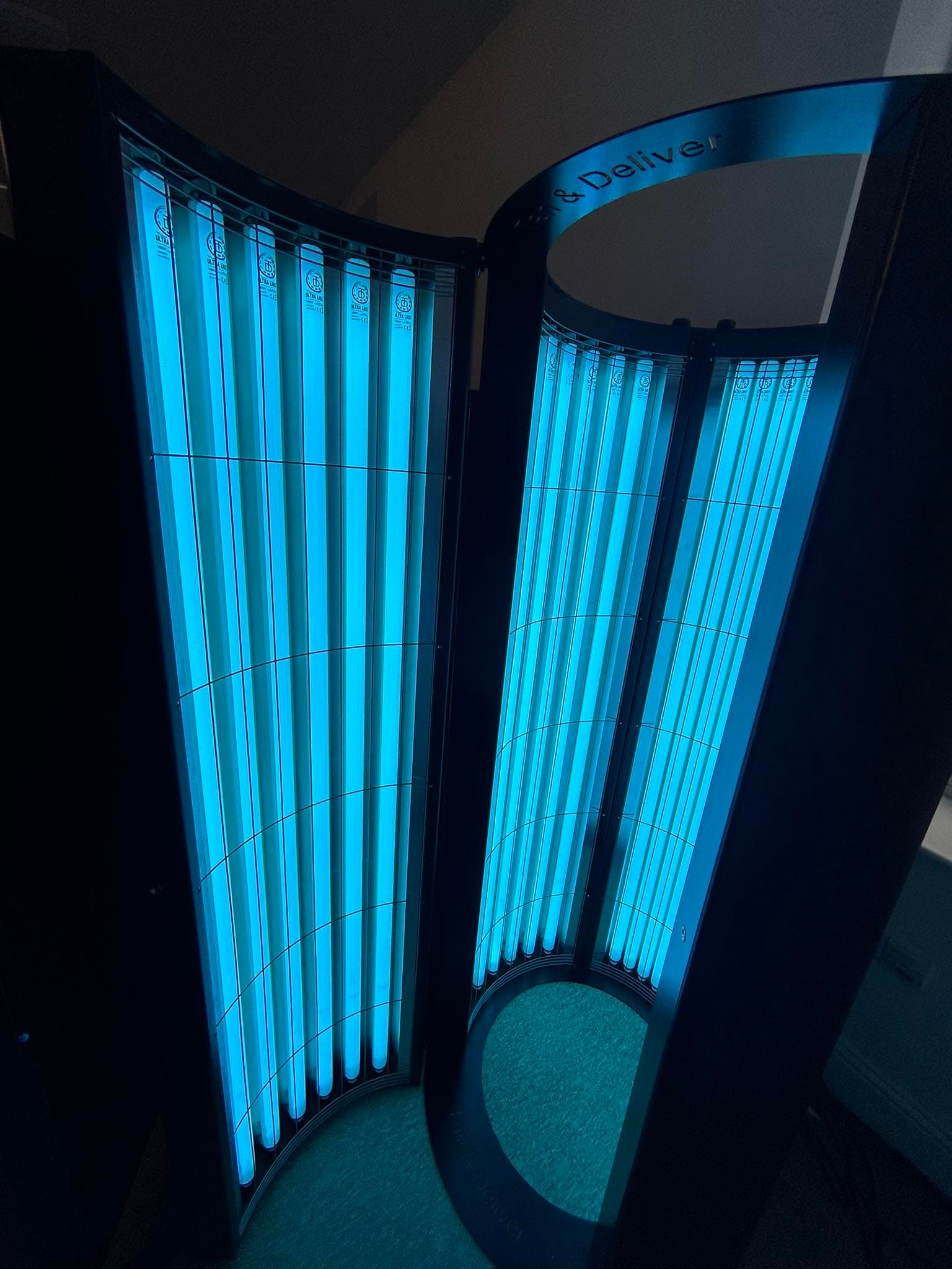 Tan & Deliver South West stand-up sunbed unit, lit from inside, showing the curved tube panels.